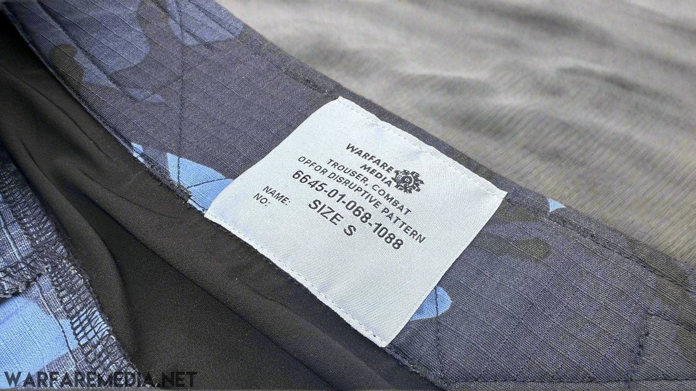 Close-up of a clothing label on dark fabric, which reads: "WARFARE MEDIA TROUSER, COMBAT, OPERATOR, OPFOR DPM TROUSERS (G3 Cut), 6654-01-086-1088, MAN NAME: _, SIZE S”. The G3 Cut Trousers blend black, gray, and shades of blue fabric and are laid on a brick surface—a staple.