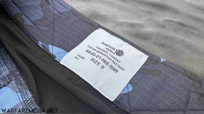 Close-up of a clothing label on dark fabric, which reads: "WARFARE MEDIA TROUSER, COMBAT, OPERATOR, OPFOR DPM TROUSERS (G3 Cut), 6654-01-086-1088, MAN NAME: _, SIZE S”. The G3 Cut Trousers blend black, gray, and shades of blue fabric and are laid on a brick surface—a staple.