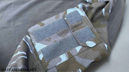 A close-up view of an OPFOR DPM UBACS (G3 Cut) sleeve by Warfare Media, featuring a mix of green and blue tones in a camouflaged pattern, with multiple Velcro patches attached to it on a grey surface. The combat shirt sleeve has various rectangular patches that suggest personalization or attachment points for other items.