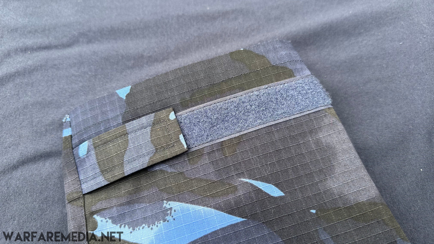 Close-up photo of a Warfare Media OPFOR DPM UBACS (G3 Cut) with a Velcro strap. The fabric features a grid texture, and the Velcro strap is prominently displayed across the top portion. The background is a plain dark surface.