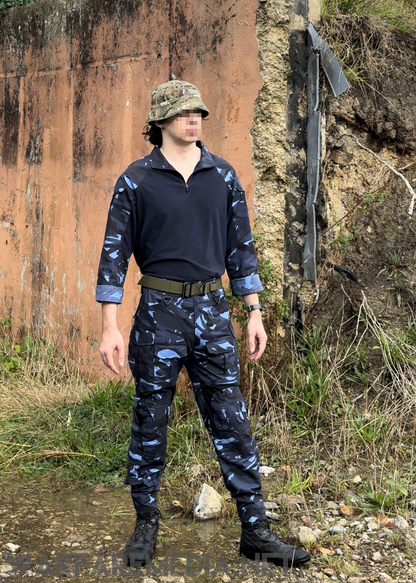 A person stands outdoors on uneven, rocky terrain in a military-inspired camo outfit from Warfare Media's OPFOR DPM full set bundle. They sport a UBACS shirt, combat trousers, and a camouflage hat. The background features a weathered wall and patches of grass. Their face is pixelated for anonymity. Pre-order now!